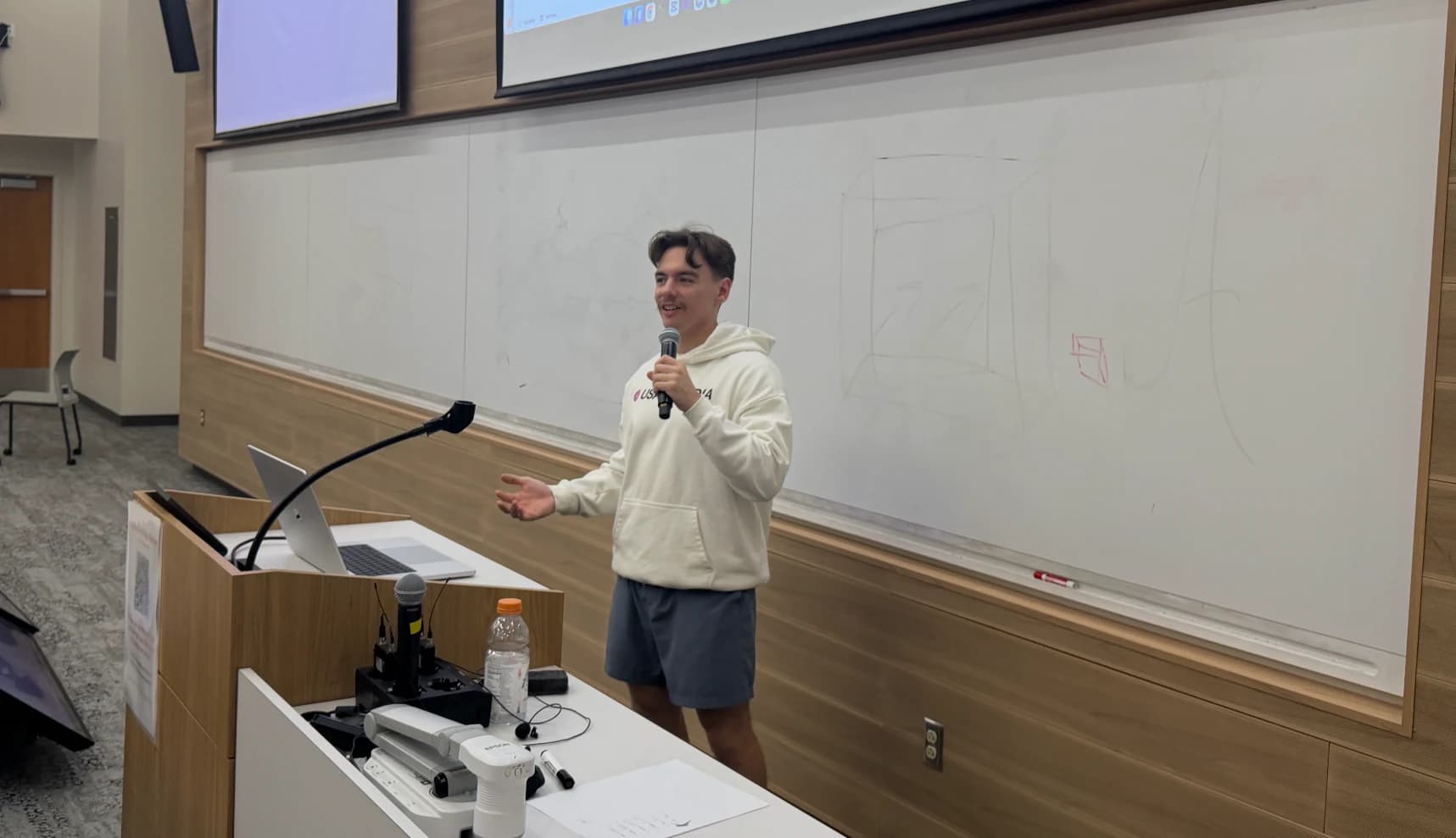 A presenter speaking with a microphone beside a whiteboard and lectern in a lecture room